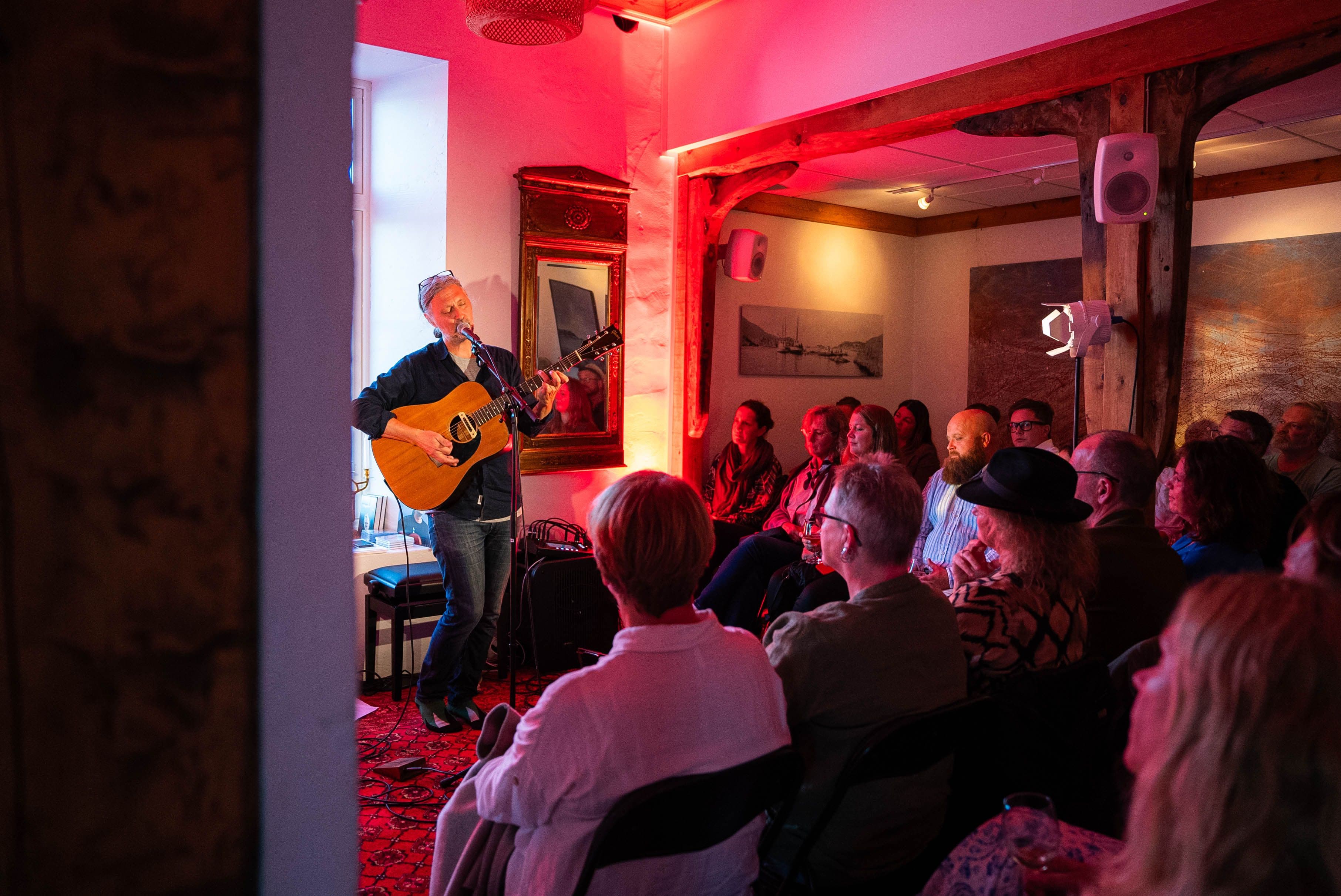 Mannlig vokalist synger og spiller gitar, omringet av publikummere i et hvitt betongrom med rødt lys.
